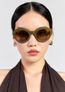 A close-up of a model wearing oversized olive green cat-eye sunglasses with dark brown lenses and small gold hoop earrings, set against a light grey background.