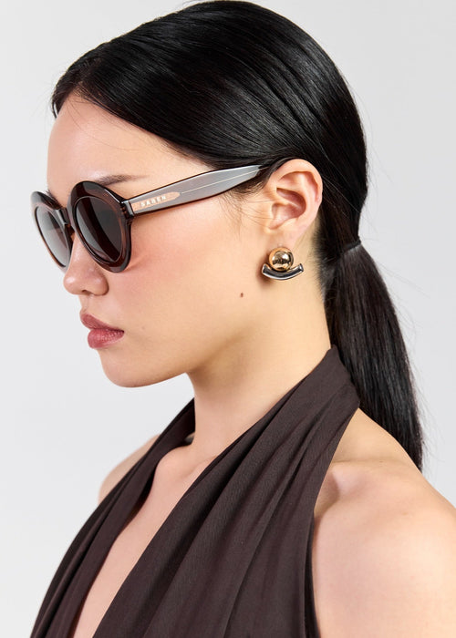 A woman in profile wearing dark cat-eye sunglasses with a grey acetate frame and gold-toned logo detailing, paired with a dark brown halt