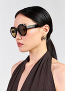 A woman in profile wearing tortoiseshell cat-eye sunglasses with gold-toned lenses and a dark brown halter top, photographed against a light grey studio background.