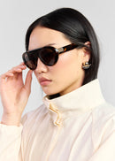 A woman with dark hair wearing aviator-style sunglasses with tortoiseshell frames and a cream high-neck blouse, set against a plain light grey background.