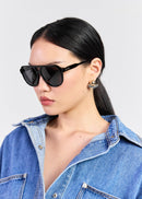 A woman with black hair pulled back wears black square sunglasses with a thin gold-tone frame and a gold-and-black earring, paired with a blue denim shirt against a light grey studio background.