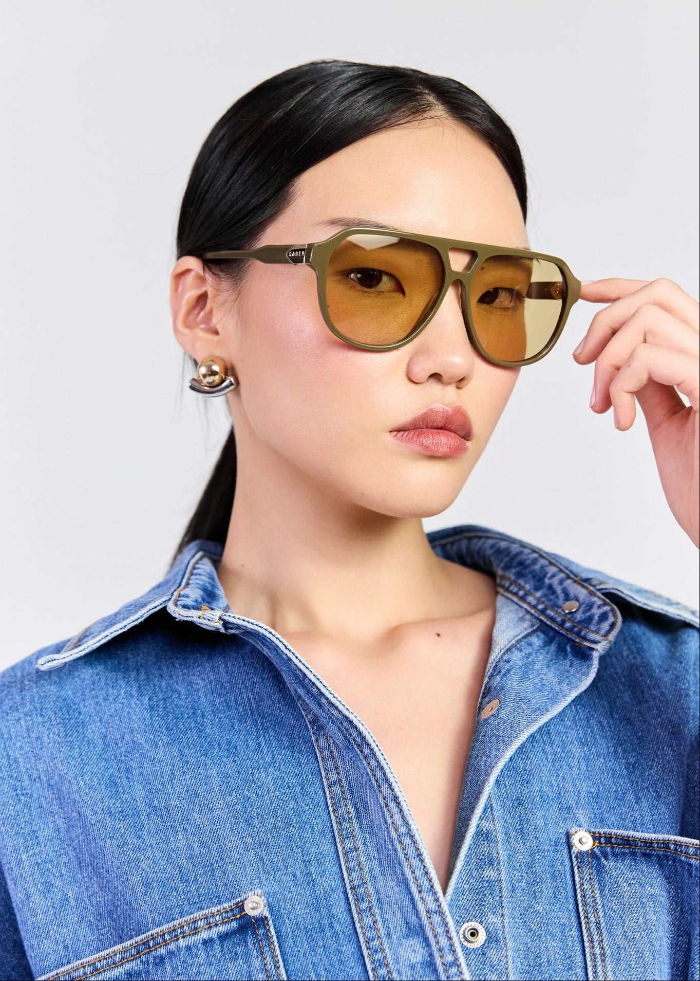 A model wearing square aviator-style sunglasses with olive green frames and amber-tinted lenses, paired with a blue denim shirt against a light grey background.