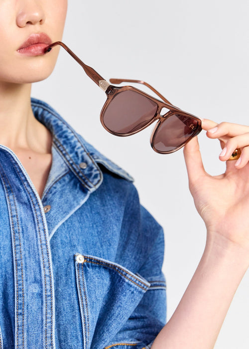 A close-up of a person wearing a blue denim shirt and holding a pair of brown sunglasses near their lips against a light grey background.