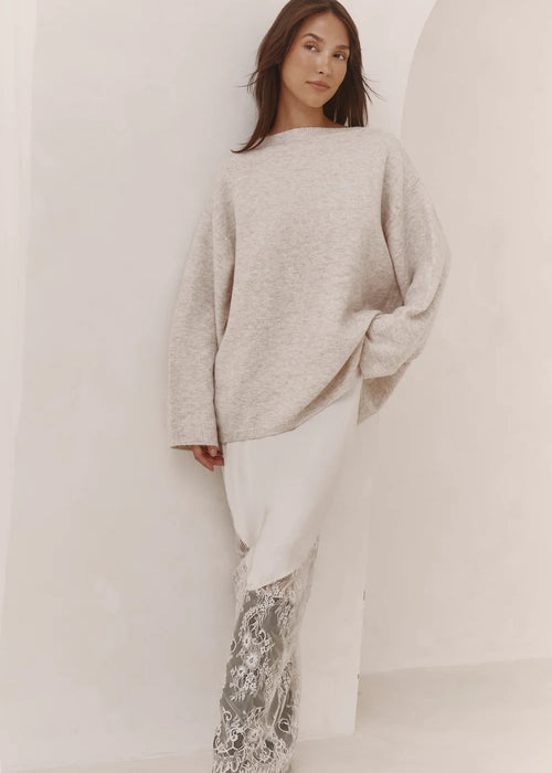A woman modelling a light beige oversized knit sweater and white pants with a silver-grey patterned lower section, standing in a minimalist white room with arched walls.