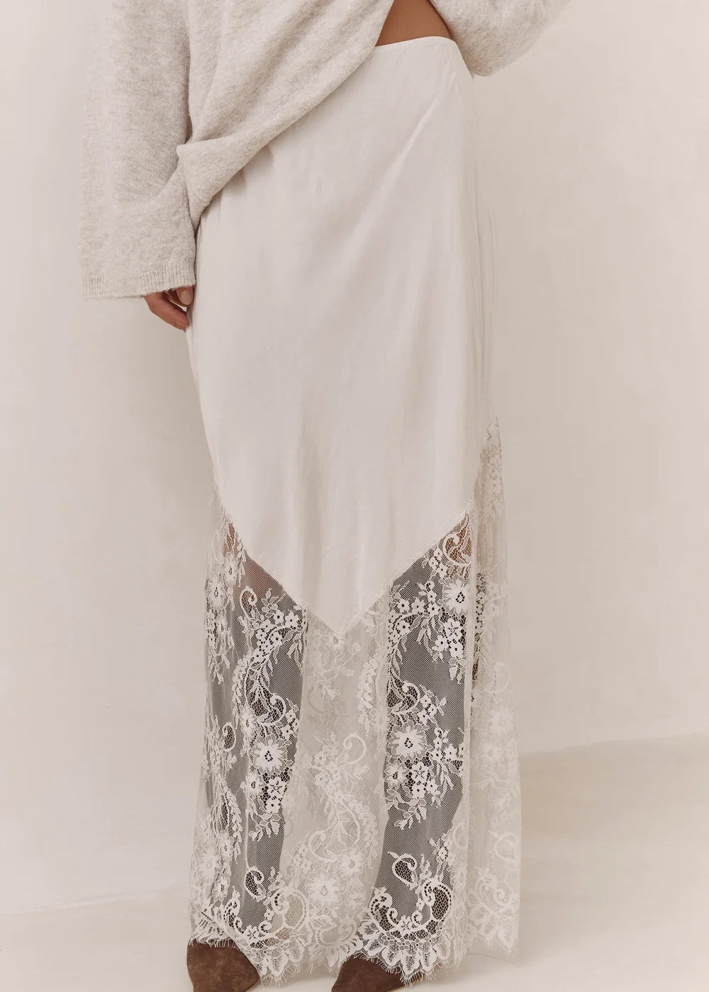A person wearing a long cream maxi skirt with a sheer floral lace overlay at the hem, paired with a light grey knit top and brown shoes, shown against a plain off-white studio background.