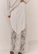 A person wearing a long cream maxi skirt with a sheer floral lace overlay at the hem, paired with a light grey knit top and brown shoes, shown against a plain off-white studio background.