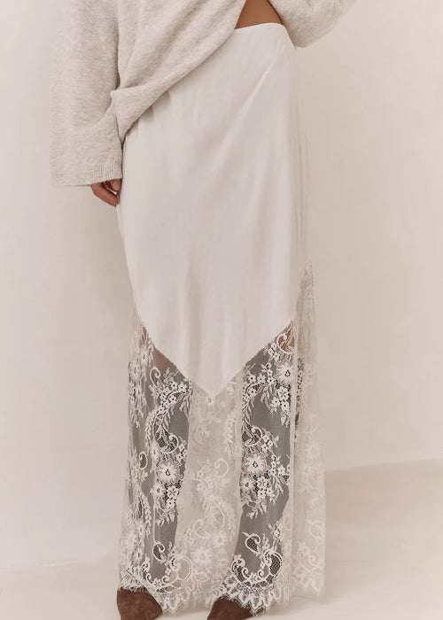 A person wearing a long cream maxi skirt with a sheer floral lace overlay at the hem, paired with a light grey knit top and brown shoes, shown against a plain off-white studio background.