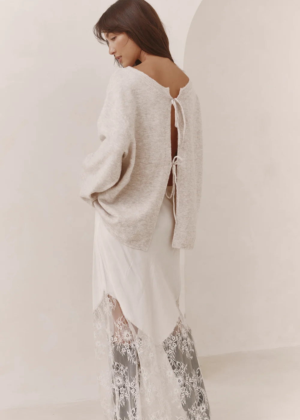 A woman wearing a cream knit cardigan over a white dress with sheer lace harem pants, photographed in a minimalist cream-colored studio setting.