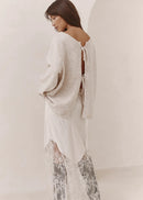 A woman wearing a cream knit cardigan over a white dress with sheer lace harem pants, photographed in a minimalist cream-colored studio setting.