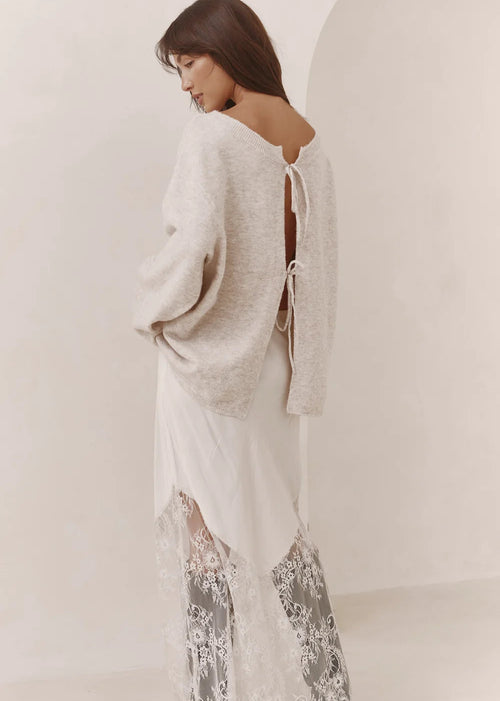 A woman wearing a cream knit cardigan over a white dress with sheer lace harem pants, photographed in a minimalist cream-colored studio setting.