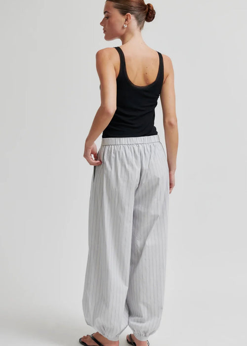 A woman modelling loose, light grey striped harem-style pants with a black tank top and black flip-flops against a plain light grey studio background.