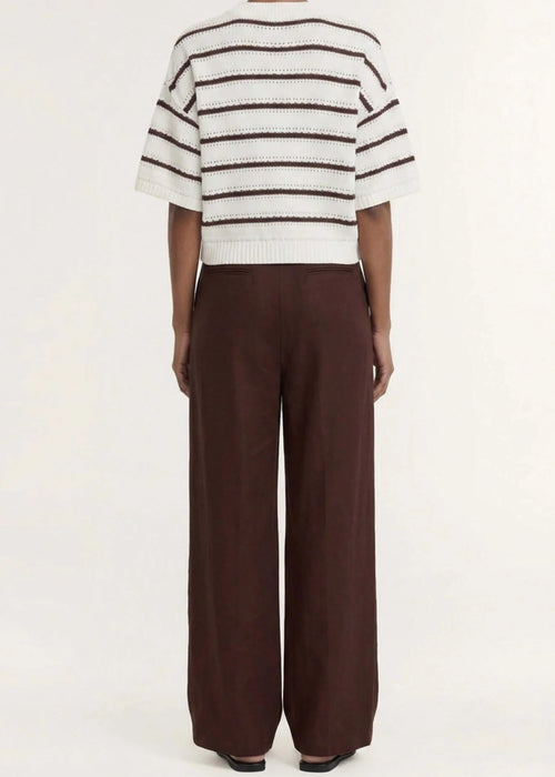 A model stands against a plain light background wearing a cream knit top with dark brown horizontal stripes, paired with dark brown wide-leg trousers and dark sandals.