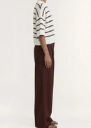 A model stands against a plain light background wearing a cream knit top with dark brown horizontal stripes, paired with dark brown wide-leg trousers and dark sandals.