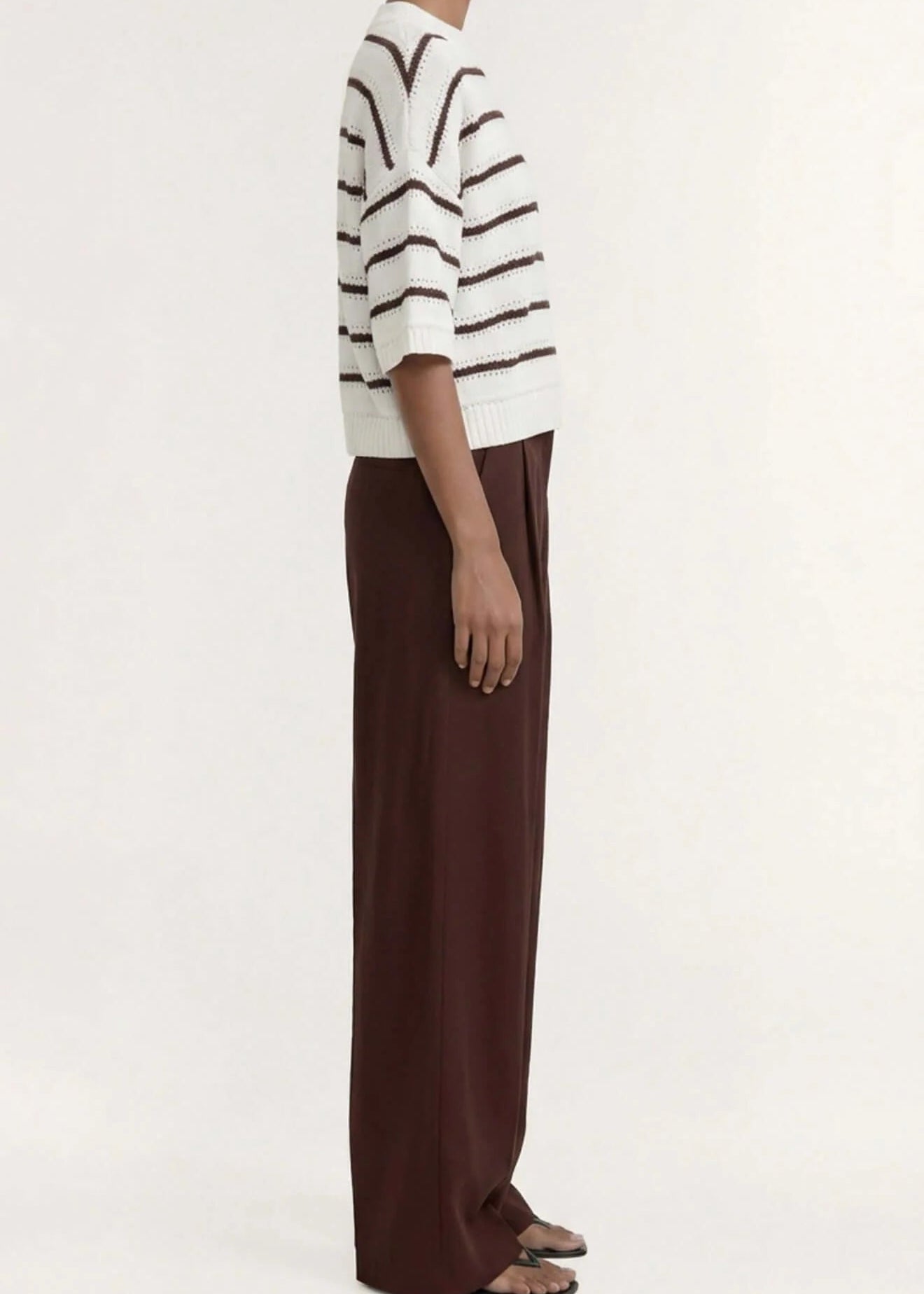 A model stands against a plain light background wearing a cream knit top with dark brown horizontal stripes, paired with dark brown wide-leg trousers and dark sandals.