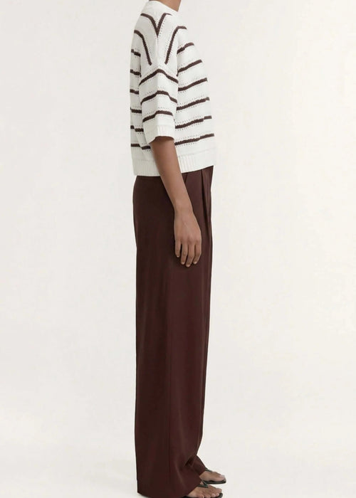 A model stands against a plain light background wearing a cream knit top with dark brown horizontal stripes, paired with dark brown wide-leg trousers and dark sandals.