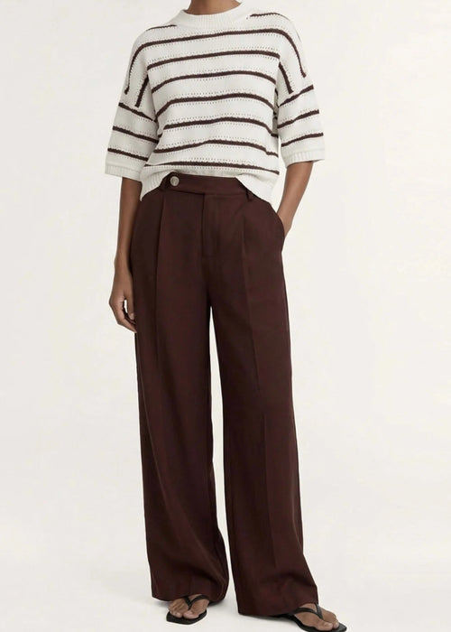 A model stands against a plain light background wearing a cream knit top with dark brown horizontal stripes, paired with dark brown wide-leg trousers and dark sandals.