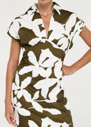 A woman viewed wearing a short-sleeve midi dress with a bold white floral print over an olive green base, set against a plain light grey studio background.