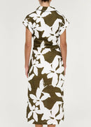 A woman viewed from the back wearing a short-sleeve midi dress with a bold white floral print over an olive green base, set against a plain light grey studio background.