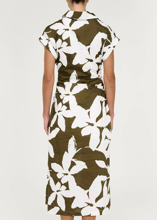 A woman viewed from the back wearing a short-sleeve midi dress with a bold white floral print over an olive green base, set against a plain light grey studio background.
