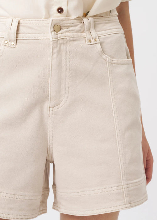 Close-up of a person wearing light beige denim shorts with a cream shirt partially visible at the top, against a plain light background.