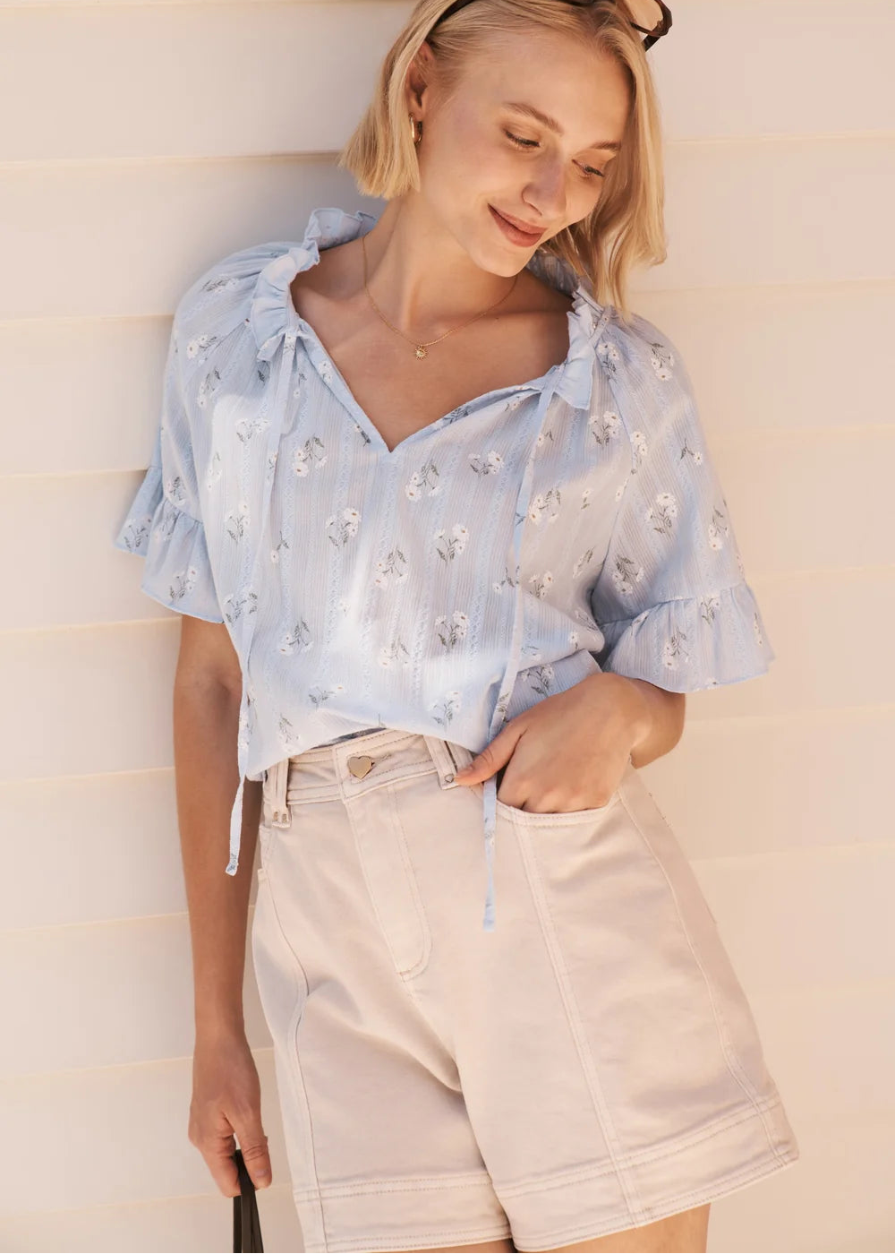 A woman wearing a light blue short-sleeve blouse with white floral embroidery and beige shorts, standing against a light beige panelled wall.