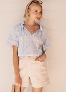 A woman modeling a light blue short-sleeve blouse with white floral embroidery and tie details, paired with cream shorts, standing against a light beige paneled wall.