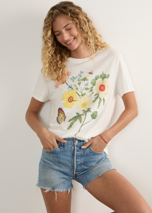 A woman stands in a studio wearing a white graphic T-shirt with a colourful floral and butterfly print, light-wash denim shorts, and white slide sandals.