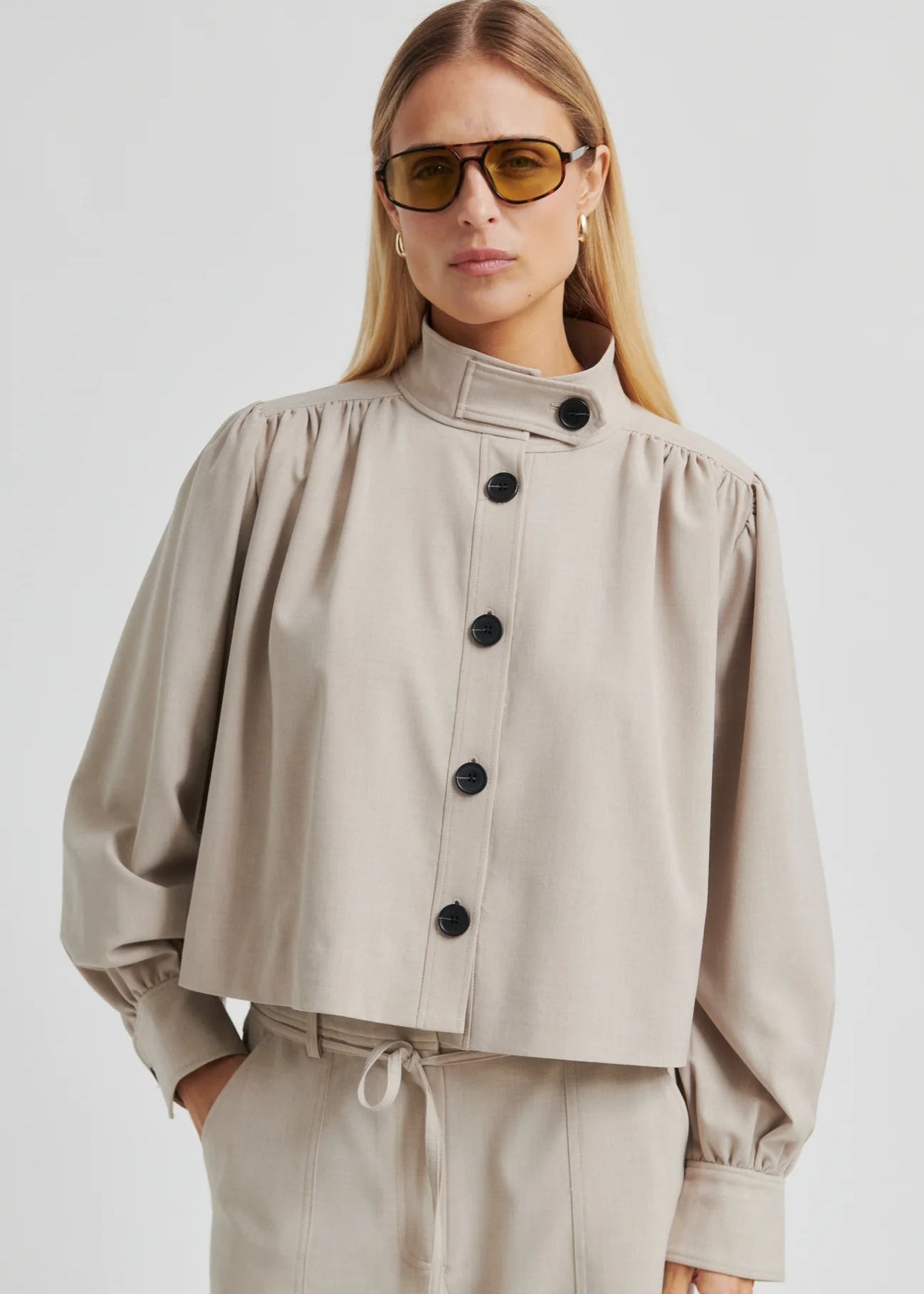 Woman wearing a beige button-up jacket and pants with sunglasses on a plain background