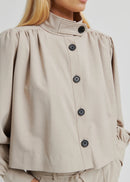 Woman wearing a beige button-up jacket and pants with sunglasses on a plain background