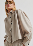 Woman wearing a beige button-up jacket and pants with sunglasses on a plain background