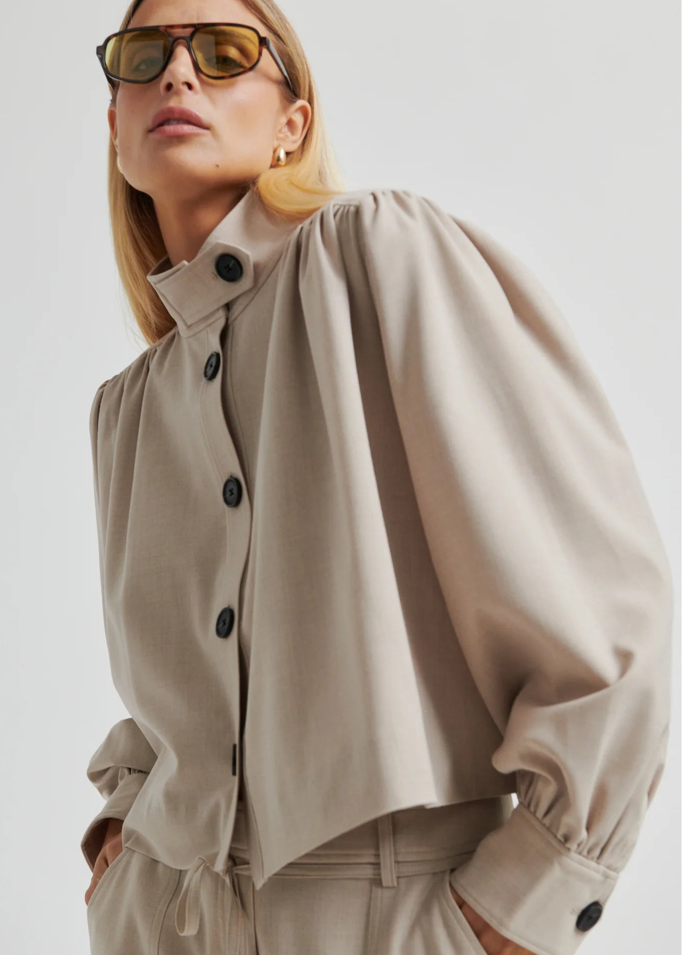 Woman wearing a beige button-up jacket and pants with sunglasses on a plain background