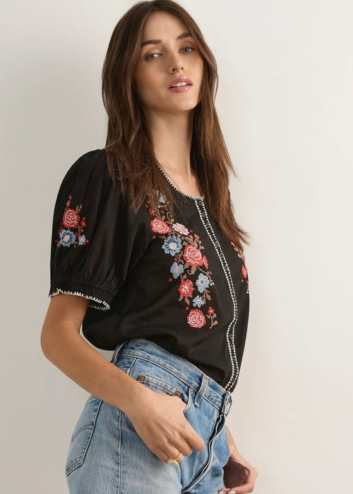 A woman modelling a black short-sleeve blouse with colourful floral embroidery on the chest and front, paired with light-wash denim shorts and brown strappy sandals, standing against a plain light grey studio background.