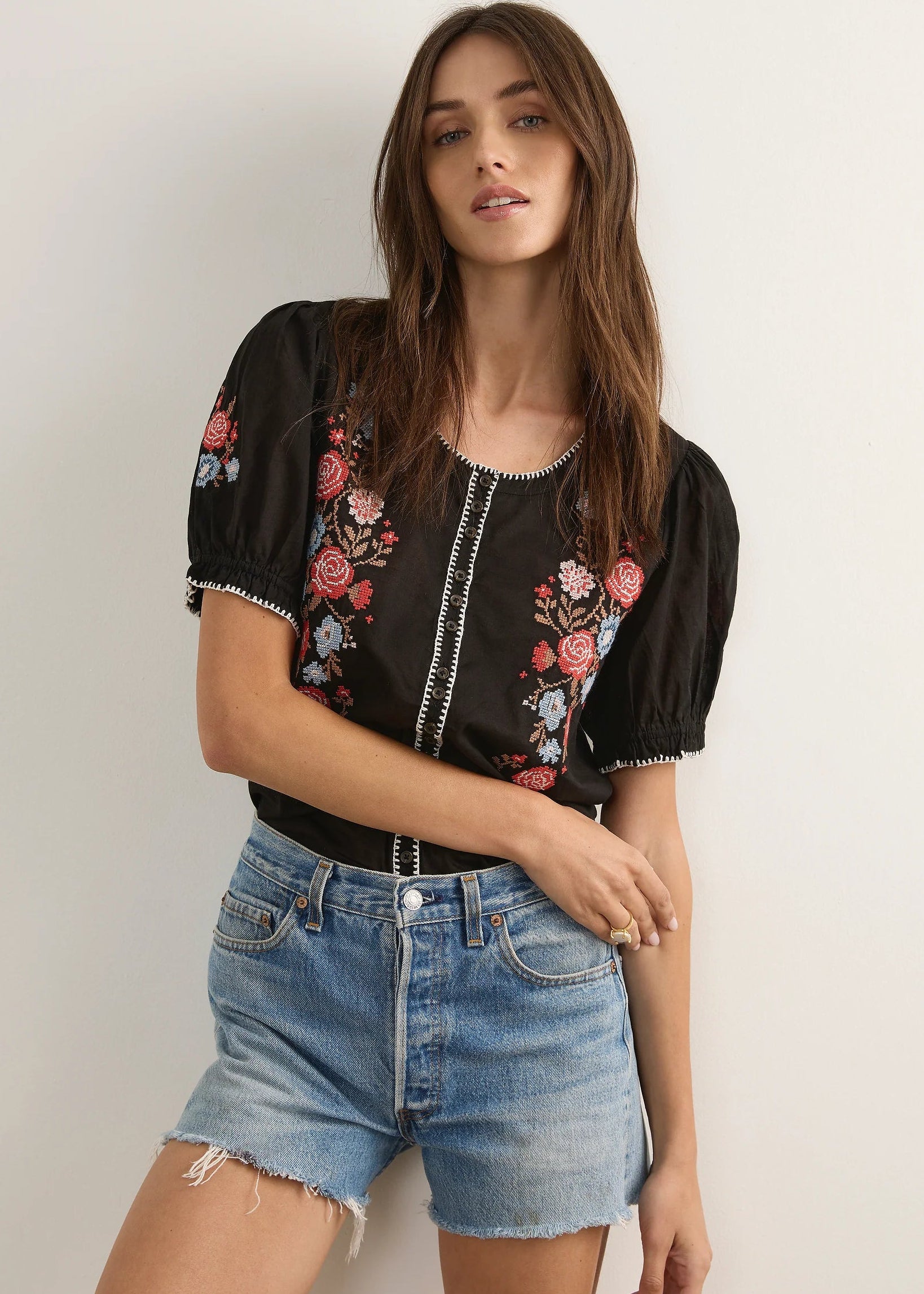 A woman modelling a black short-sleeve blouse with colourful floral embroidery on the chest and front, paired with light-wash denim shorts and brown strappy sandals, standing against a plain light grey studio background.
