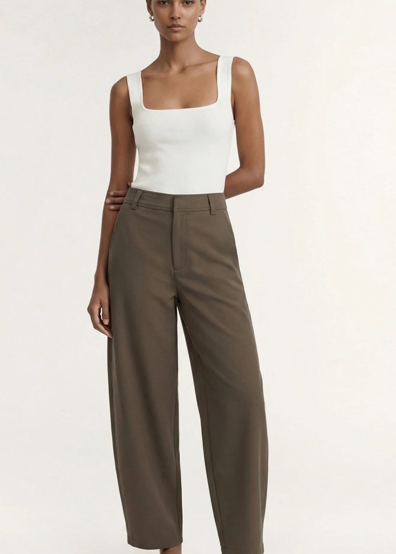 A model wearing a white sleeveless top and high-waisted, wide-leg brown trousers, paired with black flip-flops, against a plain off-white studio background.