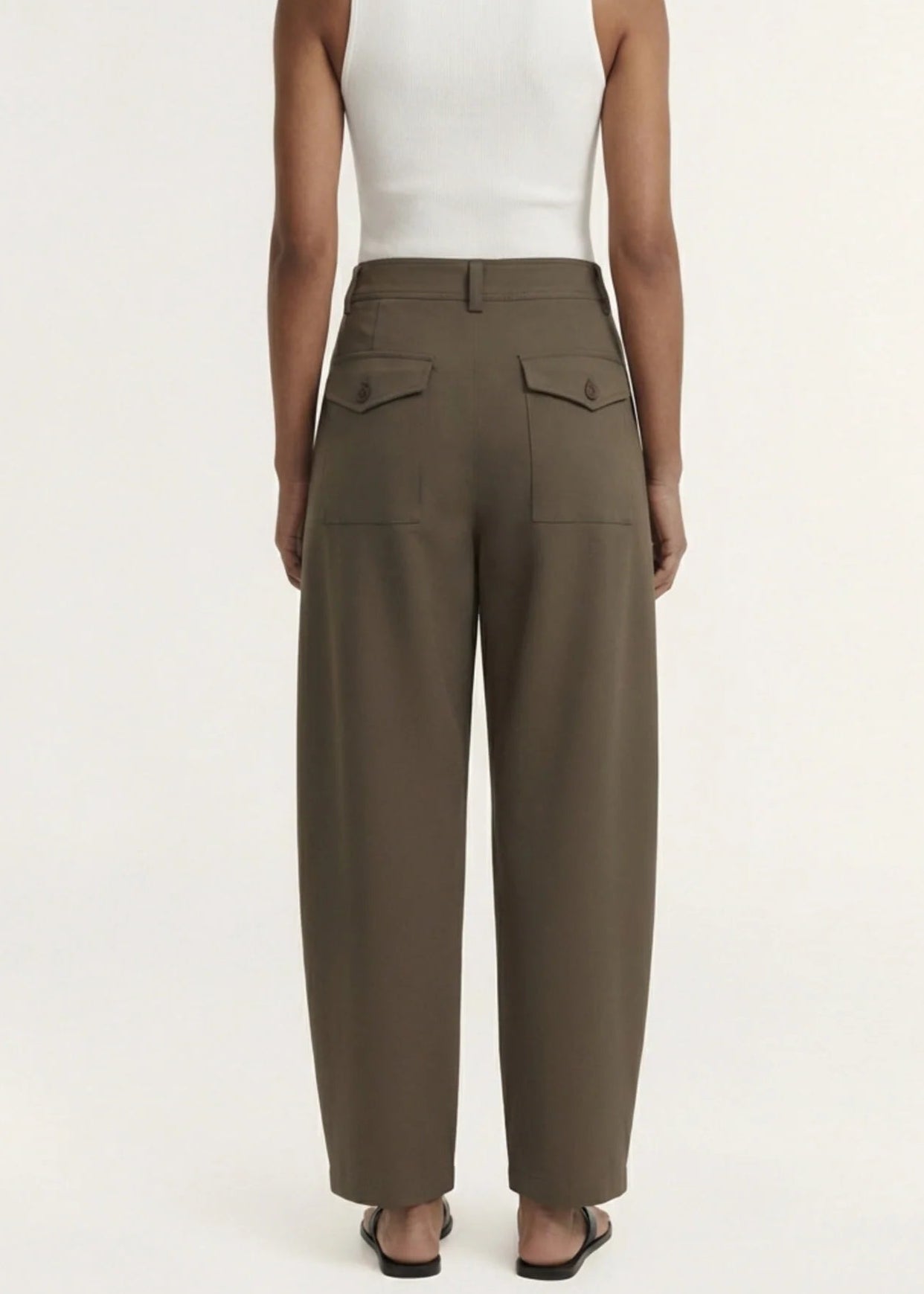 A model wearing a white sleeveless top and high-waisted, wide-leg brown trousers, paired with black flip-flops, against a plain off-white studio background.