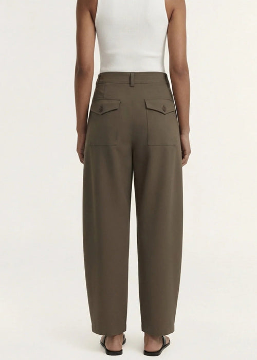 A model wearing a white sleeveless top and high-waisted, wide-leg brown trousers, paired with black flip-flops, against a plain off-white studio background.
