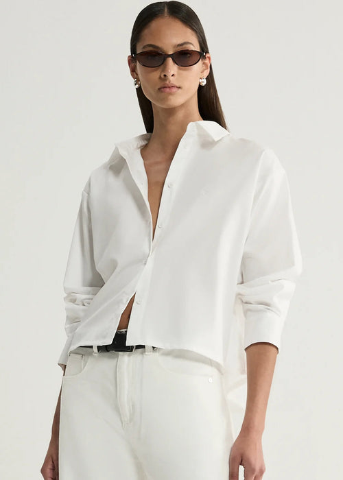 A model wearing a white long-sleeve button-up shirt with a relaxed, slightly oversized fit, paired with white trousers and dark sunglasses, against a plain light background.