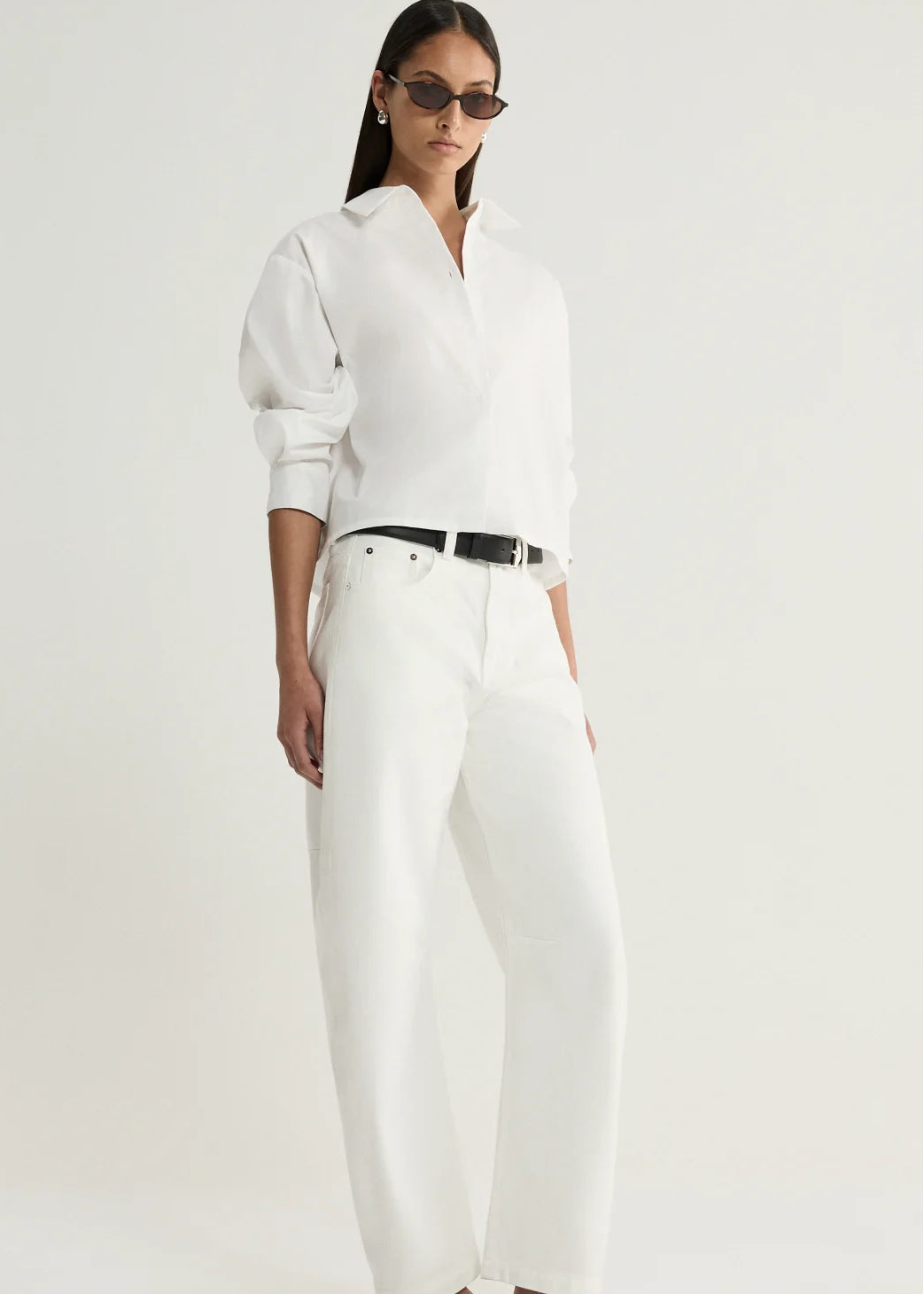 A model wearing a white long-sleeve button-up shirt, white wide-leg trousers, a black belt, and black sandals, photographed against a clean off-white studio background.