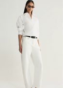 A model wearing a white long-sleeve button-up shirt, white wide-leg trousers, a black belt, and black sandals, photographed against a clean off-white studio background.