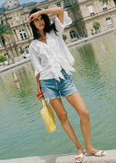 A woman stands by a reflective pond in front of a grand classical building, wearing a white ruffled long-sleeve blouse, light-wash denim shorts, white slide sandals, and holding a yellow woven handbag.