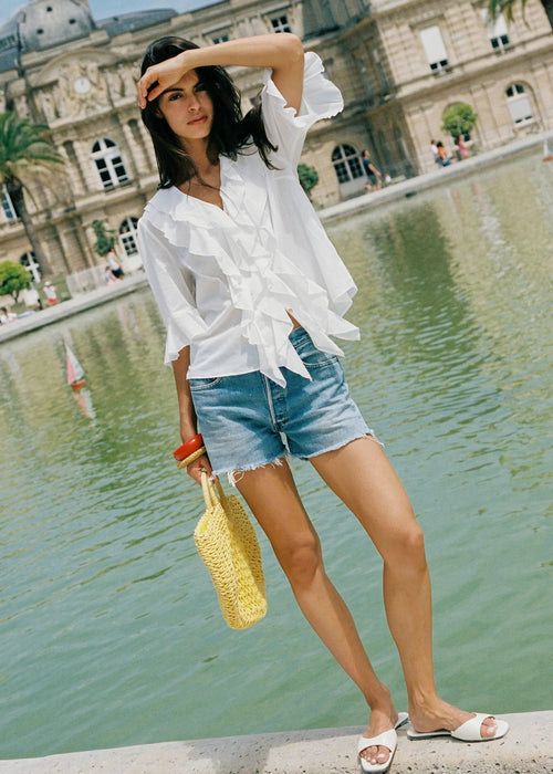 A woman stands by a reflective pond in front of a grand classical building, wearing a white ruffled long-sleeve blouse, light-wash denim shorts, white slide sandals, and holding a yellow woven handbag.
