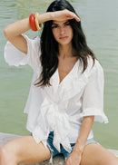 A woman sitting outdoors by the water wears a white ruffled short-sleeve blouse tied at the waist with denim shorts. She is wearing red and gold bangles and shielding her eyes from the sun with one hand.