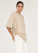 A model stands in profile wearing a loose, draped beige short-sleeve top, white shorts, and dark brown flip-flops against a plain light grey studio background.
