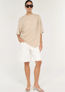 A model stands in profile wearing a loose, draped beige short-sleeve top, white shorts, and dark brown flip-flops against a plain light grey studio background.