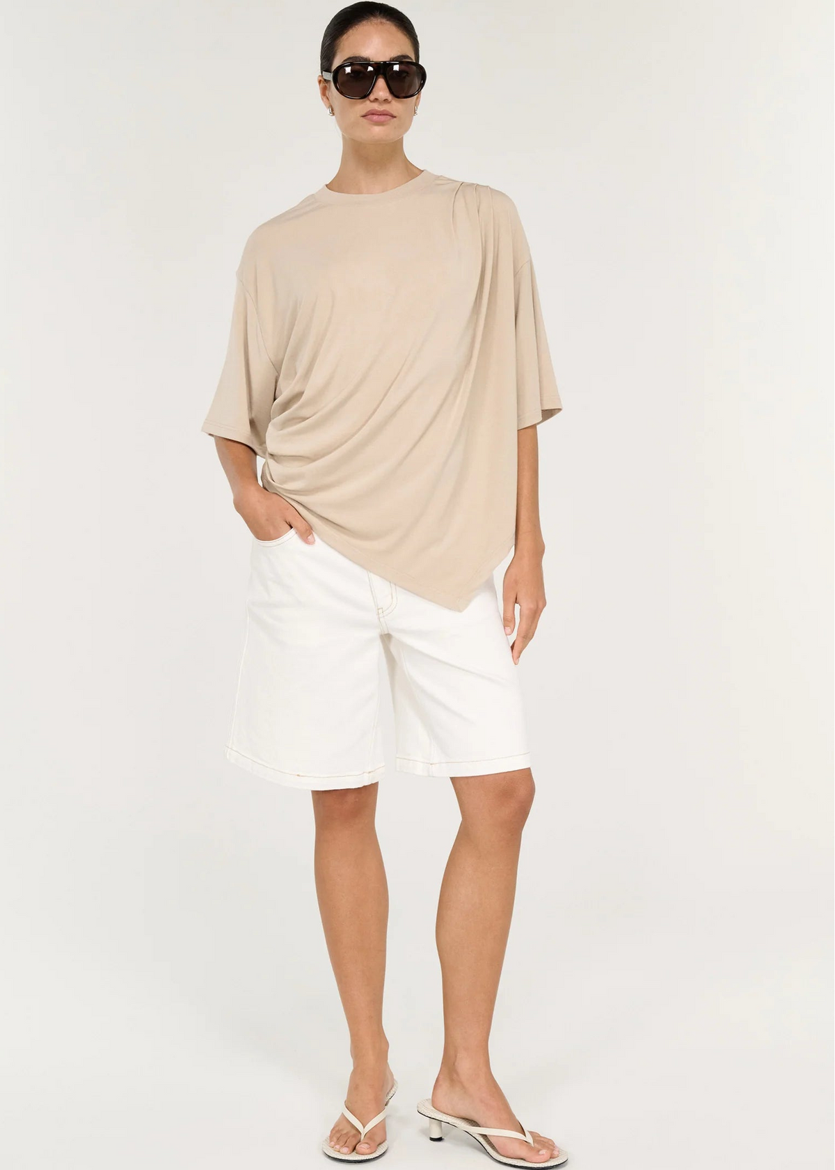 A model stands in profile wearing a loose, draped beige short-sleeve top, white shorts, and dark brown flip-flops against a plain light grey studio background.
