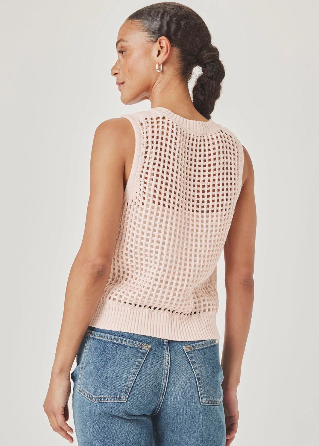 Woman wearing a light pink sleeveless knitted top and blue jeans on a plain background