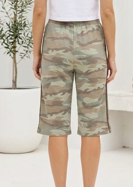A woman viewed from behind wearing camouflage knee-length shorts, a white t-shirt, and white sneakers in a bright, modern white room with a potted plant and a bench.