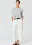 A woman modelling a light grey cardigan over a matching grey top, paired with white cropped trousers, a black belt, and black sandals, against a plain light grey studio background.