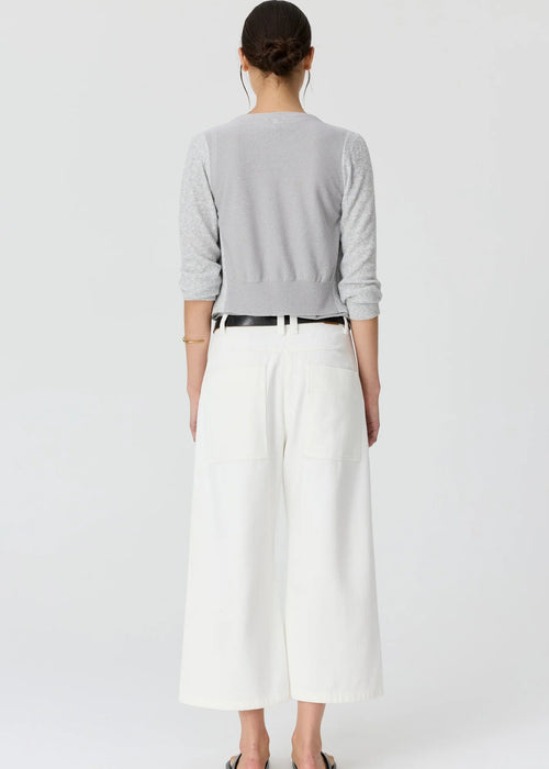 A woman modelling a light grey cardigan over a matching grey top, paired with white cropped trousers, a black belt, and black sandals, against a plain light grey studio background.