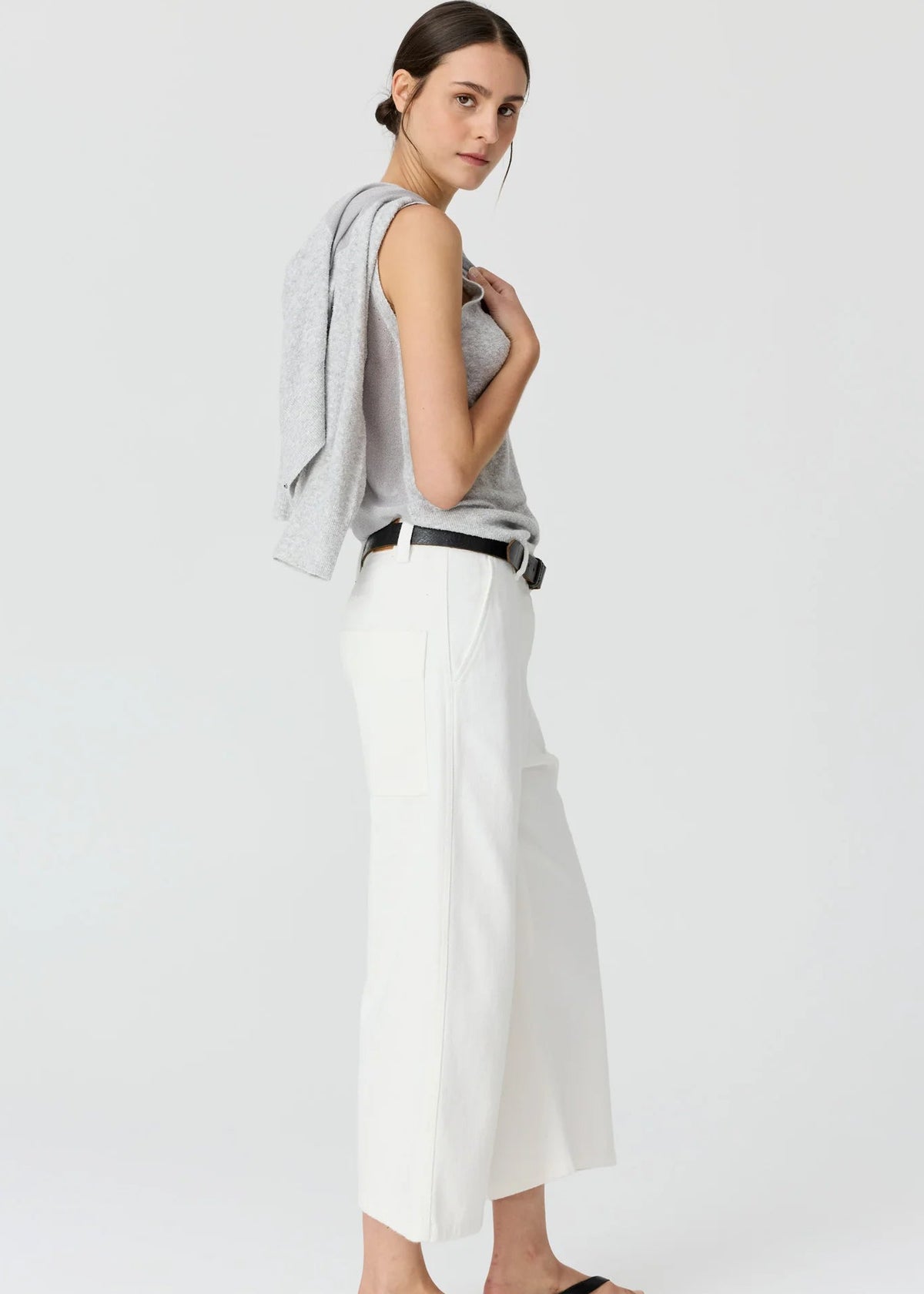 A woman modelling a light grey cardigan over a matching grey top, paired with white cropped trousers, a black belt, and black sandals, against a plain light grey studio background.